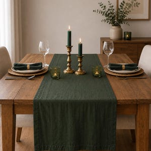 Puede incluir: Un camino de mesa rectangular de color verde oscuro hecho de tela texturizada centrado en una mesa de comedor de madera. La mesa está dispuesta con dos servicios que incluyen manteles individuales redondos de mimbre y candelabros de latón.