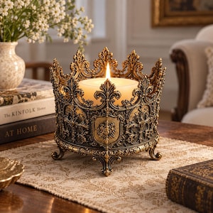 May include: A round, antique gold-toned metal candle holder shaped like a crown with intricate filigree detailing, a central shield emblem, and four curved feet. A single cream-colored pillar candle sits inside, featuring a lit flame at the center.