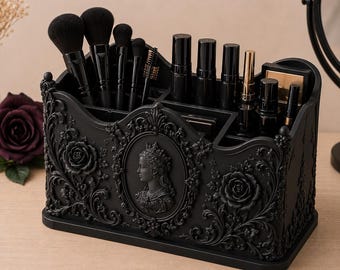 Può includere: Un organizer per trucchi rettangolare nero caratterizzato da decorazioni floreali in rilievo e un cammeo centrale raffigurante una persona con corona. L'organizer presenta diversi scomparti contenenti vari pennelli da trucco e tubetti cosmetici neri.