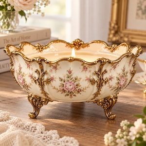 May include: A cream-colored ceramic bowl candle with a floral pattern featuring pink roses and green leaves. The bowl has a scalloped rim and ornate gold-toned metal trim and feet. A single lit wick is centered in the white wax inside the bowl.