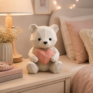 May include: A white geometric bear figurine with a faceted surface, featuring black eyes and a black nose, holding a pink geometric heart against its chest. The bear is constructed with sharp, angular planes and sits in a seated position.