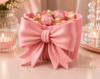 Può includere: Un contenitore rosa chiaro a forma di cupcake scanalato, caratterizzato da un grande fiocco prominente sulla parte anteriore. Il contenitore è riempito con diversi dolciumi rotondi color rosa metallizzato avvolti in carta stagnola dorata.