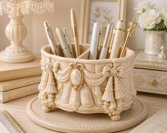 May include: A cream-colored oval desk organizer featuring ornate relief details including draped fabric swags, bows, and a central oval cameo. The container has a divided interior holding several pens and sits on a matching circular base.