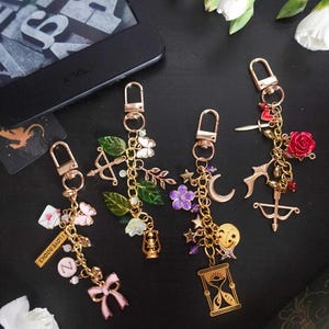 Könnte beinhalten: Vier goldfarbene Metallschlüsselanhänger mit verschiedenen Anhängern, darunter eine rosa Schleife, eine Laterne, eine Mondsichel, eine Sanduhr, eine rote Rose, ein Schwert, Pfeil und Bogen sowie ein Schild mit der Aufschrift I LOVE BOOKS.