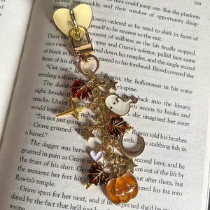 Op de afbeelding: Een goudkleurige metalen sleutelhanger met een hartvormige sluiting en meerdere bungelende bedels, waaronder een wit spookje, een oranje pompoen, een halve maan, bruine en rode herfstbladeren en kleine gouden sterretjes aan een ketting.