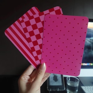 Könnte beinhalten: Drei rechteckige Karten mit abgerundeten Ecken, die fächerförmig angeordnet sind und verschiedene Muster zeigen. Die vordere Karte ist leuchtend pink mit kleinen roten Punkten, die mittlere Karte hat ein rot-pinkes Karomuster und die hintere Karte rote Streifen.