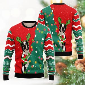 May include: A long-sleeve crewneck jumper featuring a red and green pattern with white snowflakes and wavy lines. The design displays a Boston Terrier wearing green reindeer antlers and a red, white, and green striped scarf, accented by string lights.