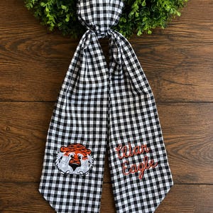 May include: A black and white gingham fabric hair scarf featuring an embroidered orange and black tiger face on one side and the embroidered red text "Cedar Eagle" on the other side, with both ends tapering to a point.