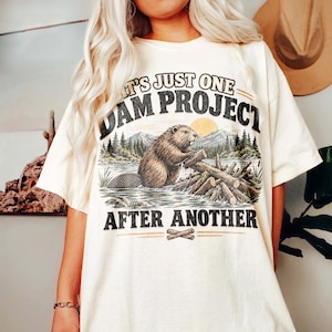Puede incluir: Una camiseta de color crema con un gráfico de un castor marrón construyendo una presa en un río con montañas y árboles de fondo. El texto dice IT'S JUST ONE DAM PROJECT AFTER ANOTHER en letras mayúsculas negras, desgastadas y en negrita.