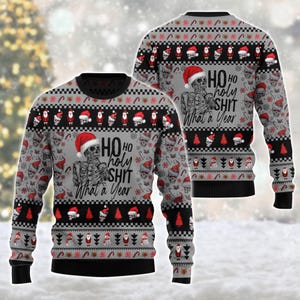 Puede incluir: Un suéter gris de manga larga con cuello redondo, puños y dobladillo negros. El diseño presenta un esqueleto con gorro de Papá Noel, sosteniendo una taza, con el texto "HO HO holy SHIT What a Year". Incluye patrones de calaveras, Papá Noel y árboles.