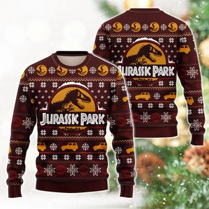 Könnte beinhalten: Ein dunkelroter Langarmpullover mit einem wiederkehrenden Muster aus weißen Schneeflocken, gelben Dinosauriersilhouetten und gelben Geländewagen. In der Mitte befindet sich das kreisförmige Jurassic Park-Logo mit einem schwarzen T-Rex.