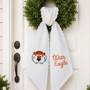 May include: A green boxwood wreath adorned with a white fabric door hanger featuring scalloped edges. The fabric displays an embroidered orange and black tiger face on the left side and the orange embroidered text War Eagle on the right side.