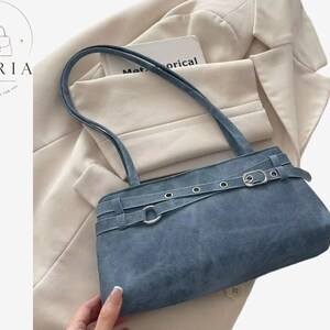 May include: A blue shoulder bag with a suede-like texture, featuring a horizontal strap across the front adorned with silver-toned metal eyelets and a buckle closure. The bag has two thin shoulder straps and a rectangular, structured silhouette.