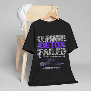 May include: A black short-sleeve t-shirt featuring a graphic print with the text DOPAMINE DETOX FAILED, REOPENED TIKTOK IN 4 MINUTES, and ALGORITHM DAMAGE. The design includes a progress bar at 0% and a purple and white glitch-style aesthetic.