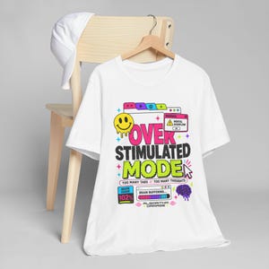 May include: A white short-sleeve t-shirt featuring a colorful graphic print with the text "OVER STIMULATED MODE", "TOO MANY TABS // TOO MANY THOUGHTS", "BRAIN USAGE 102%", "BRAIN BUFFERING", and "ALGORITHM DAMAGE", alongside a melting smiley face and brain icon.