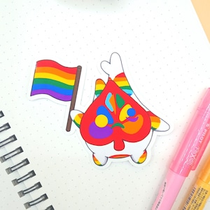 May include: A white sticker with a rainbow flag and a colorful design. The sticker is on a white notebook with a spiral binding.