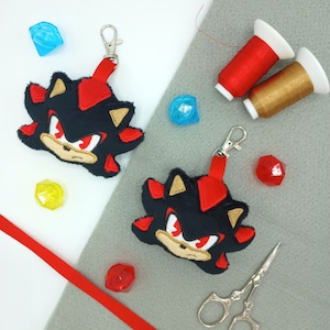 May include: Two black and red plush keychains featuring the character Shadow the Hedgehog from the Sonic the Hedgehog video game series. The keychains are on a gray surface with red and gold thread spools, blue and yellow gems, and red ribbon.