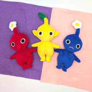 May include: Three plush toys in the shape of characters from the video game series 'NiGHTS into Dreams...' The toys are red, yellow, and blue. Each toy has a white flower on its head.