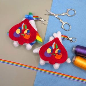 May include: Two plush heart-shaped keychains with white fabric bases and red embroidered heart patches featuring colourful abstract shapes. Each keychain includes a rainbow-striped ribbon loop with a silver metal lobster clasp attachment.