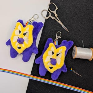 Peut inclure: Deux porte-clés en peluche violet et jaune avec des fermoirs mousquetons en métal, des ciseaux à broder argentés, une bobine de fil blanc, deux épingles à coudre et un long ruban arc-en-ciel disposés sur une surface noire et blanche.