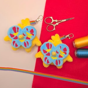 May include: Two yellow plush bird keychains with blue butterfly-shaped wings featuring pink and yellow patterns, attached to rainbow-striped ribbon clips. Also included are silver metal embroidery scissors, one yellow thread spool, one blue thread spool.