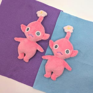 Peut inclure: Deux jouets en peluche roses avec des accents de fleurs sur la tête. Les jouets ont de grands yeux et une expression triste. Les jouets sont assis sur un fond violet et bleu.