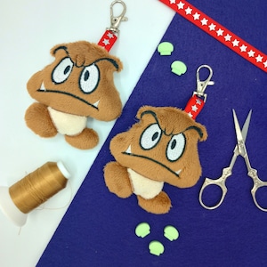 Może przedstawiać: Dwie brązowe pluszowe figurki breloczków przedstawiające postać z gry wideo z ponurym wyrazem twarzy. Figurki są przymocowane do czerwono-białych breloczków w kształcie gwiazd. Figurki znajdują się na niebiesko-białej powierzchni z zielonymi guzikami w kształcie grzybów.