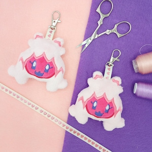 May include: Two pink and white plush keychain charms shaped like a cartoon character with a pink face and a red mouth. The charms are hanging from silver keychains.