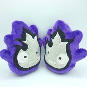 May include: Two plush purple mittens with a flame-like design in black and white. Each mitten features a light gray face with black eyes and a smiling mouth. The mittens are soft and appear to be made of a plush material.