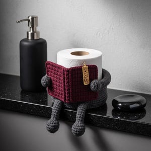 May include: A dark gray crocheted toilet paper holder shaped like a figure sitting and reading a dark red crocheted book. The figure has dangling legs and arms holding the book, which features a small gold-toned rectangular metal bookmark charm.