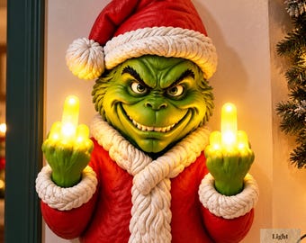 Op de afbeelding: Een groene wanddecoratie van een figuur met een rode kerstmuts en jas met witte randen. Het figuur heeft een ondeugende uitdrukking en steekt beide middelvingers op, die verlicht zijn met heldere gele lampjes van binnenuit.