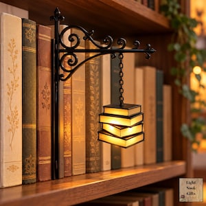 May include: A black metal wall-mounted bracket with a decorative scroll design supports a hanging light fixture shaped like a stack of three illuminated books. The light emits a warm glow from between the book covers, suspended by a chain.