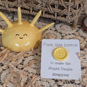 May include: A yellow ceramic sun-shaped figure with a smiling face and a white card featuring a yellow button and the text: Push this button to make the Stupid People disappear. The card is square with black printed lettering and a glossy finish.