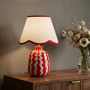 May include: A table lamp with a red and white wavy striped ceramic base and a white lampshade featuring a scalloped edge trimmed in red. The lamp emits a warm glow, illuminating the surface it rests upon next to a clear glass vase of flowers.