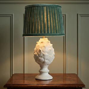May include: A table lamp featuring a white ceramic base sculpted in the shape of an artichoke. The lamp is topped with a pleated, dark teal velvet lampshade. The light emits a warm glow from beneath the shade, illuminating the ceramic petals.