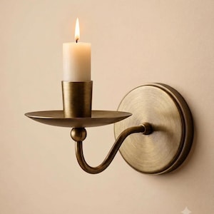 May include: A wall-mounted candle sconce made of brushed brass with a circular backplate, a curved arm, and a flat circular drip tray. A single white pillar candle sits in the center of the tray with a lit flame burning at the top.