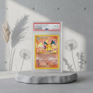 Op de afbeelding: Een 1999 Pokemon Game Charizard Holo ruilkaart beoordeeld als GEM MT, ingesloten in een doorzichtige plastic beschermhoes. De kaart toont het oranje Charizard-personage, 120 HP en tekst inclusief Charizard, Flame Pokemon en Fire Spin aanvalsdetails.