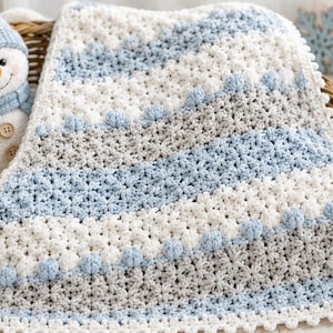 May include: A crocheted baby blanket featuring horizontal stripes in white, light blue, and soft gray. The blanket has a textured star-like stitch pattern and a decorative scalloped border. It is folded inside a woven wicker basket.
