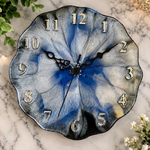 May include: A round wall clock with a scalloped edge featuring a swirling resin design in shades of deep blue, silver, and black. The clock face displays raised silver-colored numerals from 1 to 12 and includes black ornate hour and minute hands.
