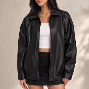 May include: A black long-sleeve jacket with a collar and a front zipper closure, worn over a white sleeveless top and a black skirt. The jacket features a smooth finish, long sleeves with cuffs, and an elasticized hemline at the waist.