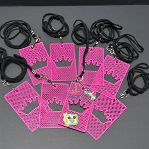 May include: A collection of seven rectangular hot pink plastic badge holders, each featuring a die-cut crown shape at the top and a textured mesh pattern. Each holder is attached to a black cord lanyard with a metal swivel hook clasp.