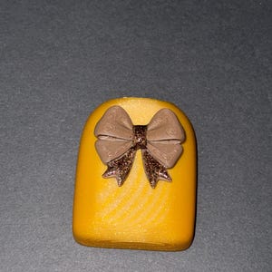 May include: A bright yellow, rounded rectangular object featuring a textured surface. Centered on the front is a three-dimensional bow in a muted brown shade, with the center knot and the bottom tails accented with a dark brown glitter finish.