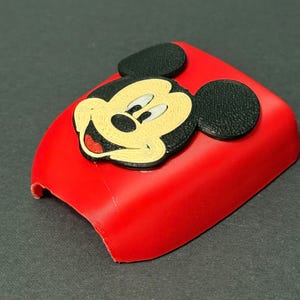 May include: A bright red, curved plastic cover featuring a raised, textured emblem of Mickey Mouse. The character has black ears, a cream-colored face, white eyes, and a red mouth. The object has a smooth, matte finish and a hollow, arched shape.
