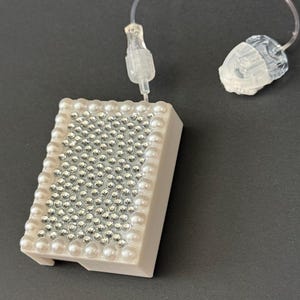 May include: A rectangular medical device pump cover decorated with a grid of silver rhinestones and a border of white faux pearls. The device is connected by a thin, clear plastic tube to a translucent medical infusion set component.
