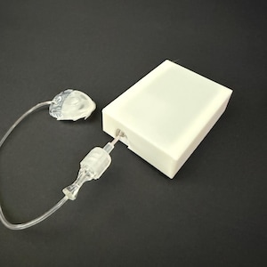 May include: A white rectangular plastic housing unit connected by a thin, clear flexible tube to a small, translucent, textured component. The tube features a clear cylindrical connector piece positioned between the housing and the end component.