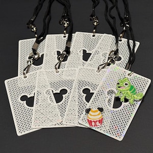 May include: A collection of white rectangular lanyards featuring a repeating textured pattern and a central cutout shaped like a mouse head. One lanyard displays a green chameleon enamel pin and a colorful cupcake enamel pin with mouse ears.
