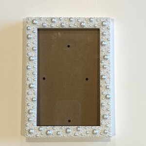 May include: A rectangular picture frame featuring a white border densely decorated with an assortment of white and silver-toned faux pearls of varying sizes, surrounding a central rectangular opening with a brown backing board.