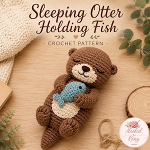 Könnte beinhalten: Ein braun-cremefarbenes Häkelotter-Spielzeug, das einen hellblauen Fisch hält. Der Text lautet Sleeping Otter Holding Fish CROCHET PATTERN. Der Otter hat geschlossene Augen, eine kleine schwarze Nase und einen gestickten Mund. Ein handgefertigtes Stofftier.