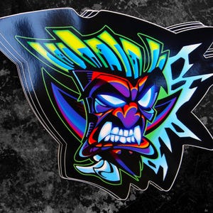 May include: A colorful sticker featuring a stylized, angry-looking face with sharp teeth and a blue, green, and yellow mohawk. The face is surrounded by a black and white lightning bolt design.
