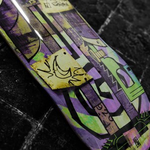 May include: A purple, green, and yellow skateboard deck with a graphic design featuring abstract shapes and a small yellow sticker with a crescent moon and a bird design.